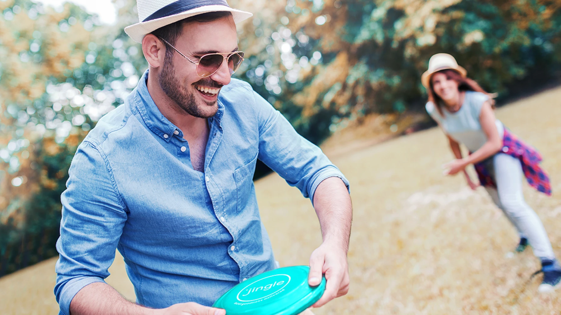 Man wearing sunglasses and a hat smiling while preparing to throw a teal frisbee that says "jingle," with a woman in the background smiling and ready to catch, playing together in a sunny park.