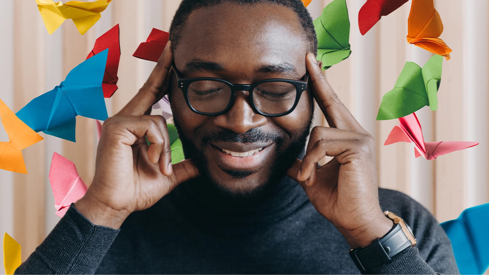 A smiling man with glasses and a beard touches his temples while keeping his eyes closed, surrounded by colorful origami butterflies, symbolizing creativity or imagination.