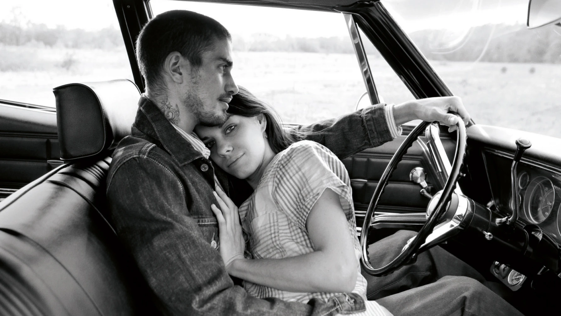 Black-and-white photo of a couple sitting in a vintage car, with the woman resting her head on the man's chest while he drives, capturing an intimate and serene moment.