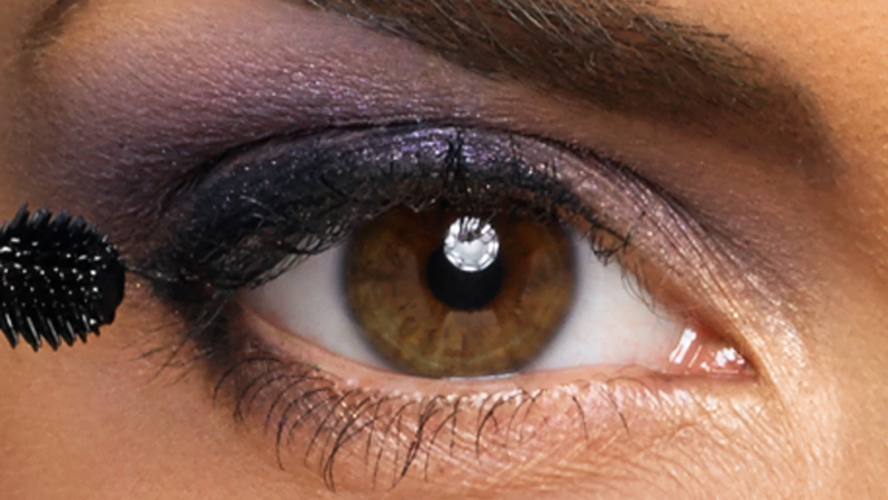 Close-up image of a person's eye with brown iris, wearing dark purple and black eyeshadow. A mascara wand is applying mascara to the upper eyelashes. The skin tone is warm, and the eyebrow is well-groomed.