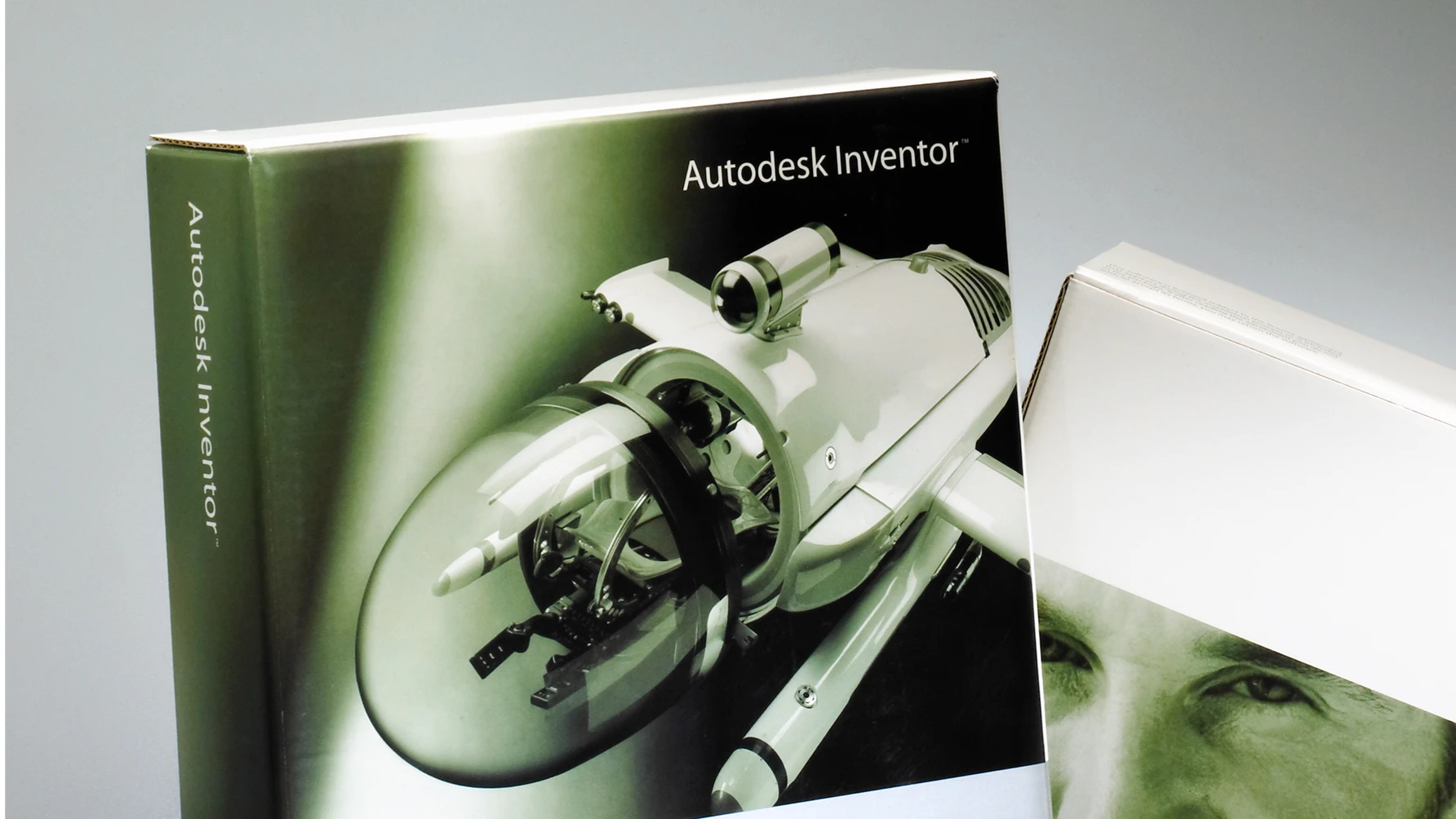 Product packaging for Autodesk Inventor software, featuring a stylized 3D rendering of a futuristic, compact aircraft or drone on the front cover, with sleek white bodywork and a transparent cockpit. The background is a smooth gradient, and the side of the box is labeled "Autodesk Inventor." Another box with a partial image of a man's face is slightly visible beside it.