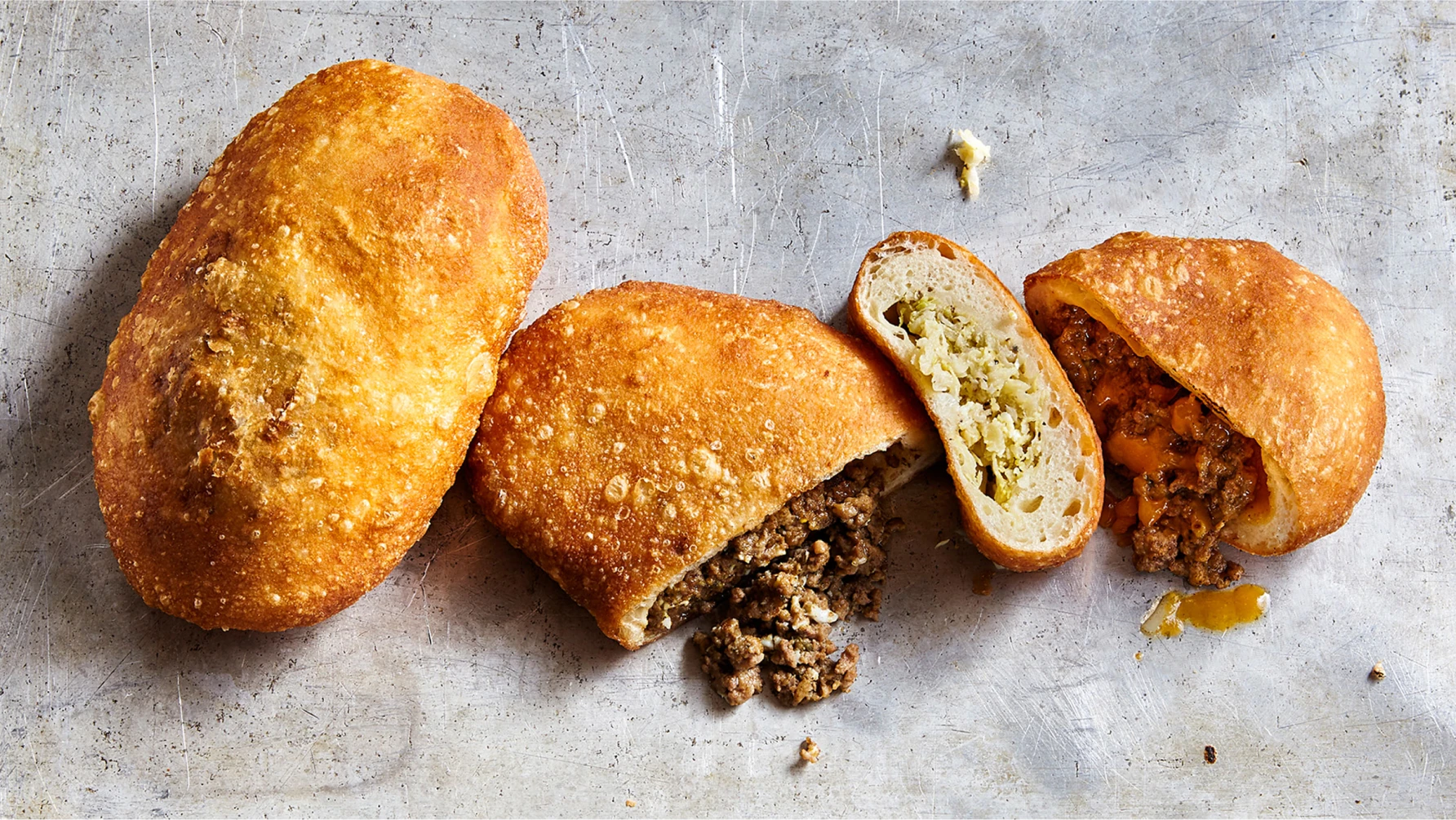 Four golden-brown fried buns displayed on a scratched metal surface. Two of the buns are whole, while the other two are cut open to reveal different savory fillings: one with ground meat, another with sauerkraut, and the last with a meat and cheese mixture. Crumbs and small bits of filling are scattered around them.