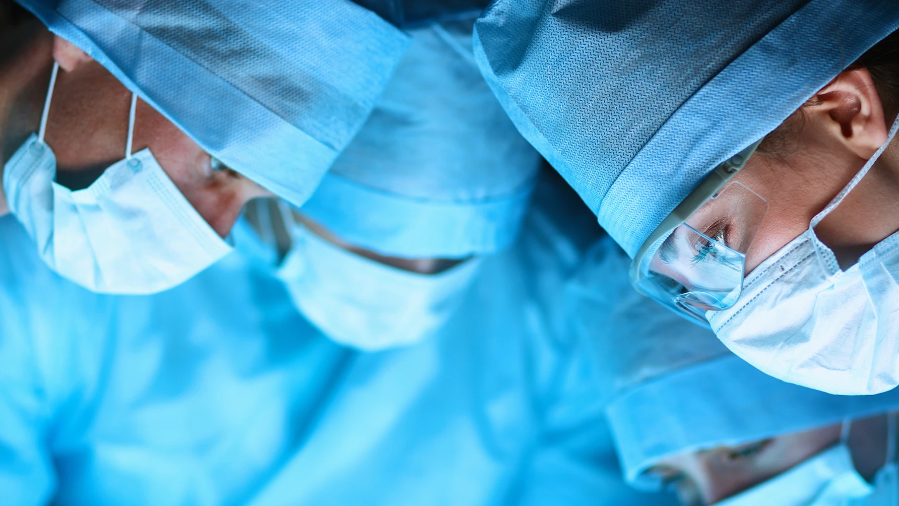A close-up view of a surgical team wearing blue scrubs, surgical masks, caps, and protective eyewear, intently focused on a medical procedure under bright operating room lights.