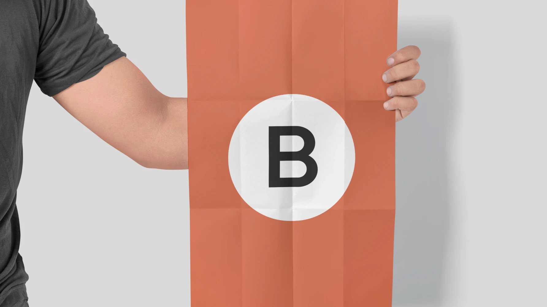 A person wearing a dark gray t-shirt holds a vertical, folded orange poster. In the center of the poster is a large white circle containing a bold, black letter "B". The background is a light gray, and the person is cropped at the shoulders, focusing attention on the poster design.