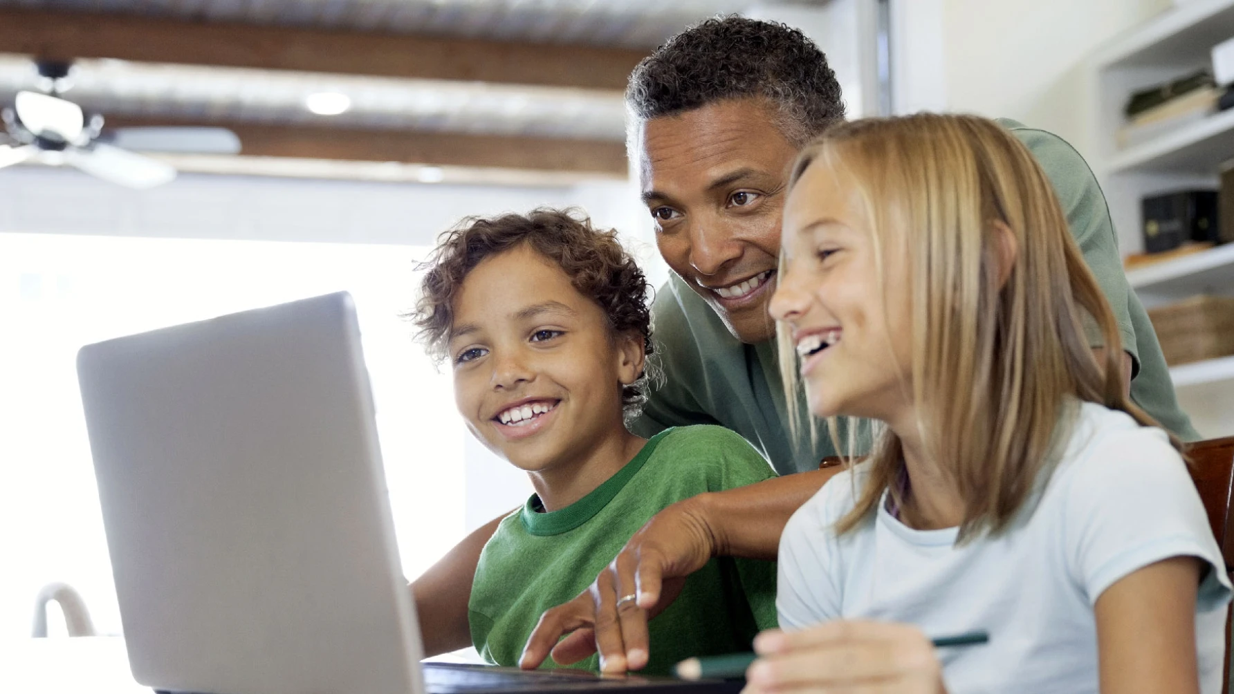 A smiling man and two children, a boy and a girl, gathered around a laptop at a table. They appear engaged and happy, suggesting a shared learning or entertainment experience in a bright, cozy home setting.