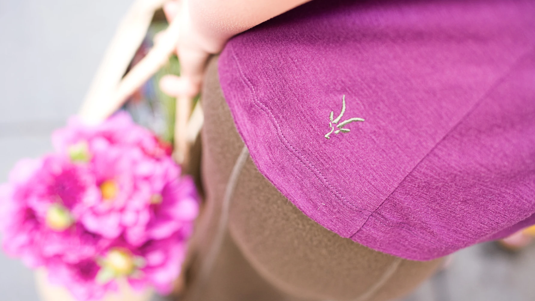 Close-up of a person wearing a magenta shirt with a small green embroidered logo near the hem. The person is holding a woven basket with vibrant pink flowers, viewed from an overhead perspective. The background is softly blurred.