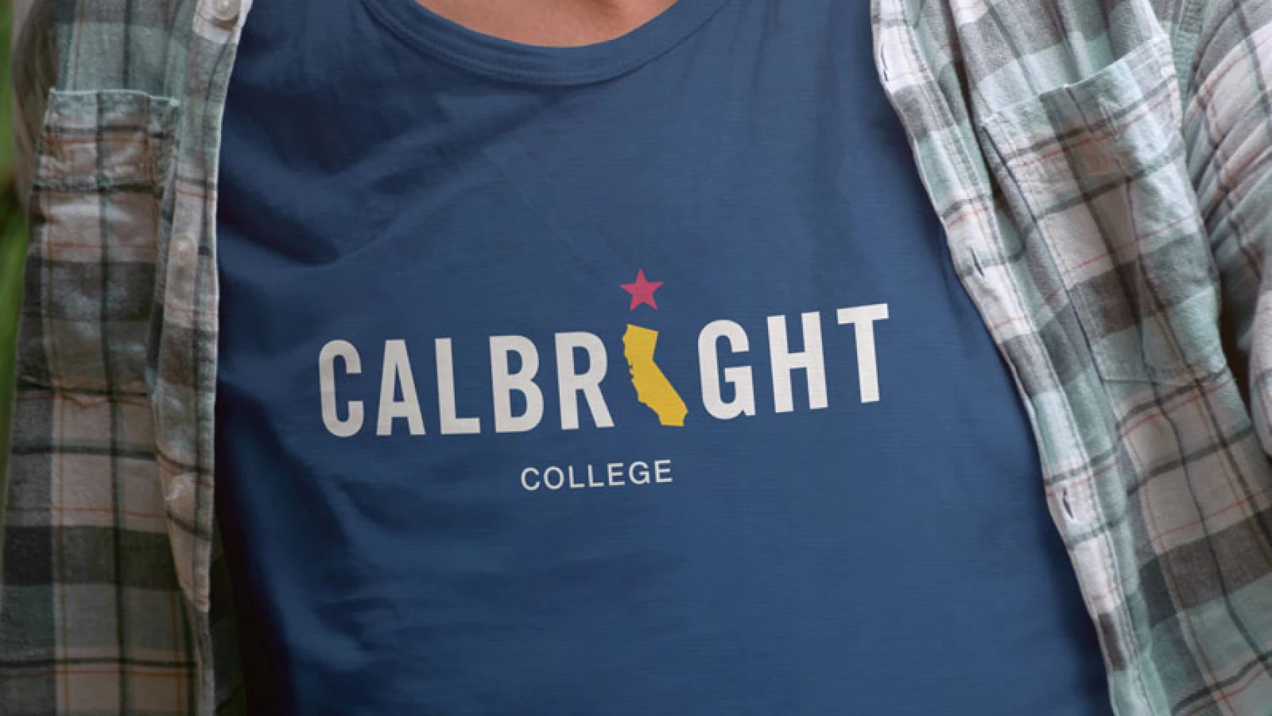 Close-up of a person wearing a blue T-shirt with the text “Calbright College.” The design features the word “CALBRIGHT” in capital letters, with the letter "I" replaced by a yellow silhouette of the state of California and a red star above it. The person is also wearing a plaid shirt over the T-shirt.
