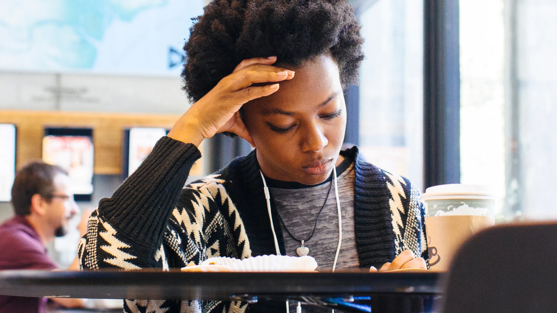A young woman with curly hair is sitting at a table in a cafe, wearing a patterned sweater and earphones. She appears focused and deep in thought while studying or reading, with a cup of coffee and a pastry in front of her.