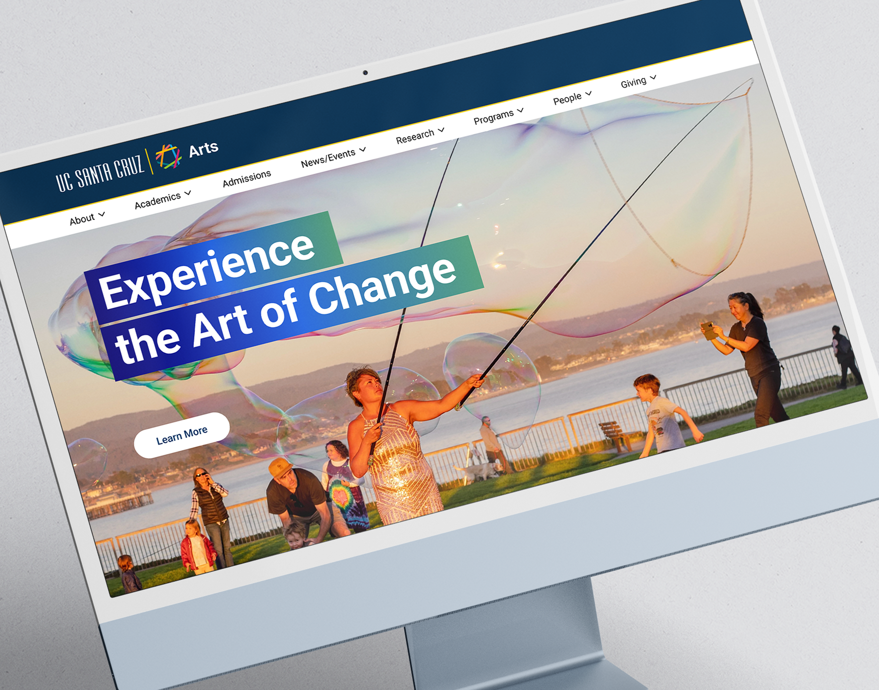 Computer screen displaying the UC Santa Cruz Arts website homepage. The main banner shows people making giant soap bubbles near a waterfront at sunset. Overlay text reads “Experience the Art of Change” with a “Learn More” button beneath. The website menu includes links for About, Academics, Admissions, News/Events, Research, Programs, People, and Giving.