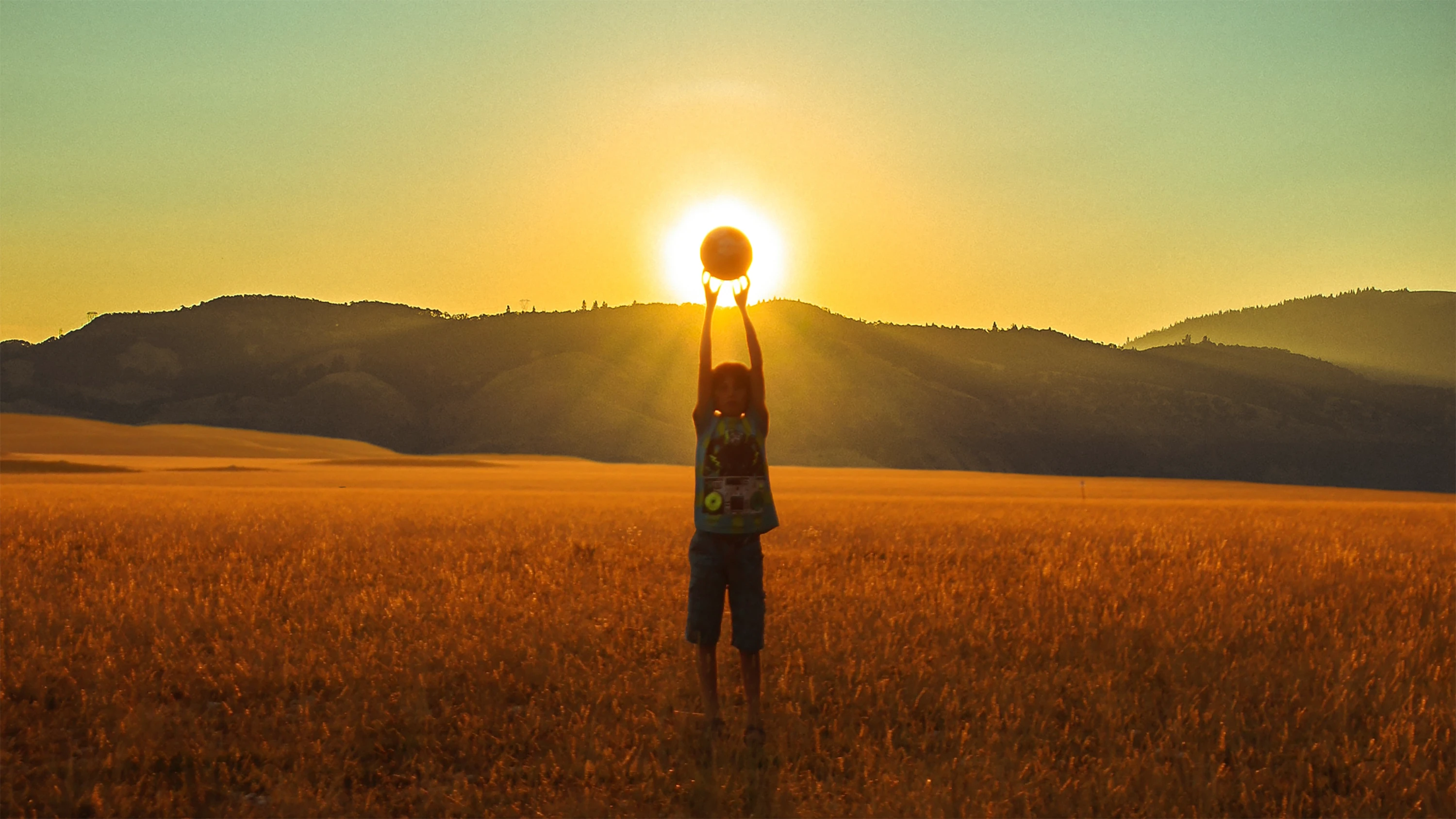 A child standing in a golden field at sunset, holding a ball above their head in perfect alignment with the sun, creating the illusion that they are holding the sun itself, with silhouetted hills in the background.