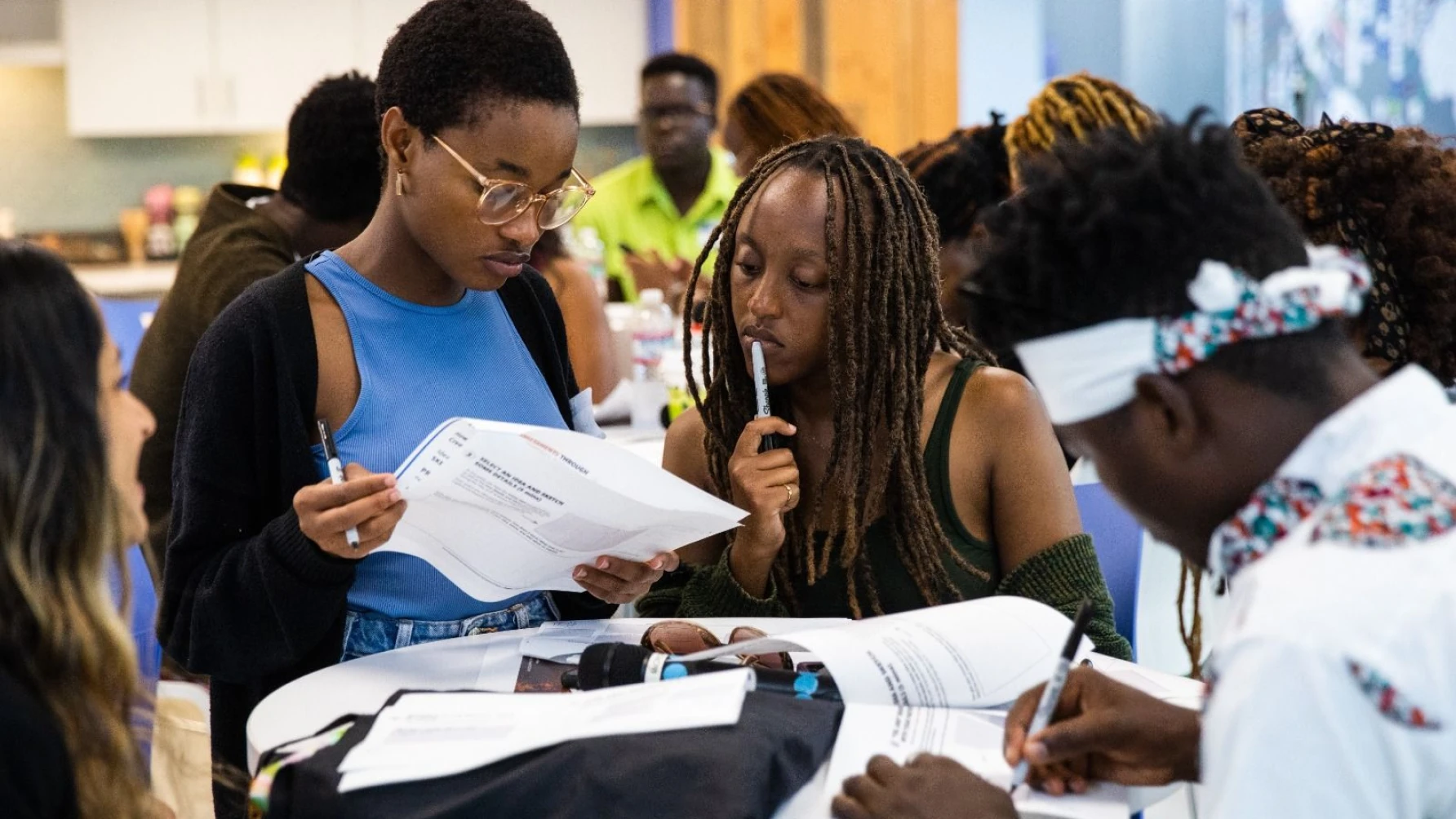 A group of young adults engaged in a collaborative learning session around a table. Two individuals in the center are closely reviewing a printed worksheet, pens in hand, while others around them read or write. The atmosphere appears thoughtful and focused, with papers, notebooks, and materials spread across the table. The setting is a modern, brightly lit room with a diverse group of participants.