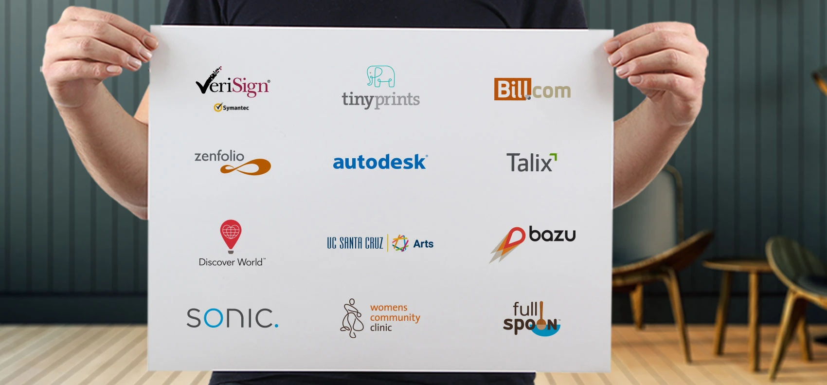 A person holds a white poster board featuring a grid of various company and organization logos. These include VeriSign, TinyPrints, Bill.com, Zenfolio, Autodesk, Talix, Discover World, UC Santa Cruz Arts, Bazu, Sonic, Women’s Community Clinic, and Full Spoon. Each logo is distinct in color and style, representing diverse industries from technology and finance to healthcare and education. The background setting is a modern room with wooden floors and mid-century chairs, blurred out to focus attention on the logos.