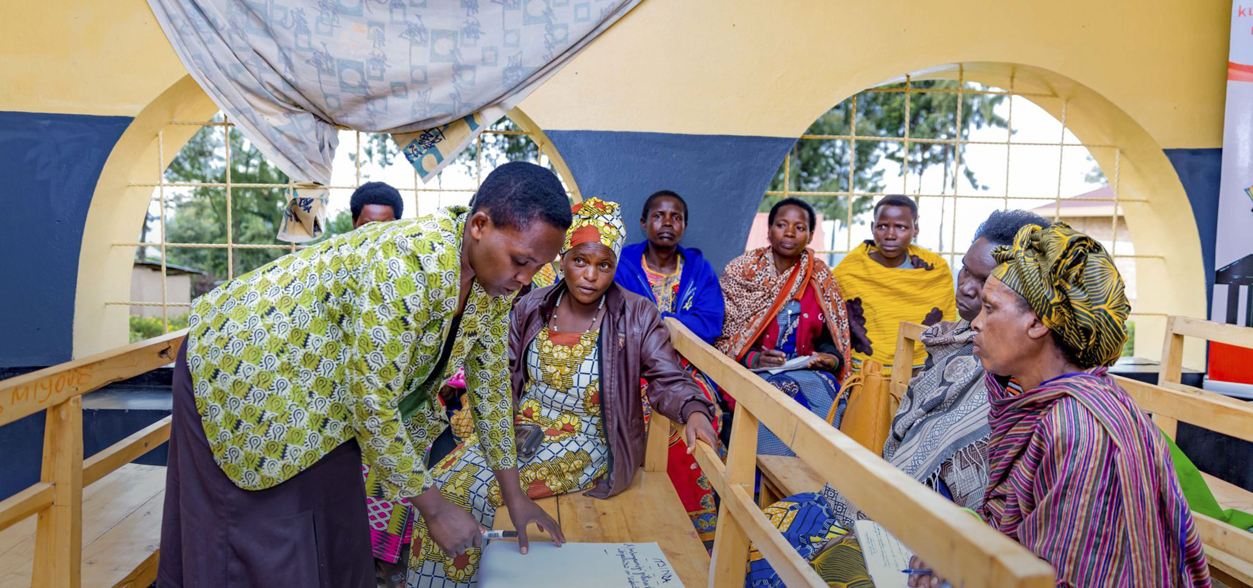 A group of women, dressed in colorful traditional African attire, sit together in a semi-circle inside a bright room with arched windows. One woman stands and writes on a piece of paper on the table, while others attentively observe and engage in the session, suggesting a community meeting or workshop.
