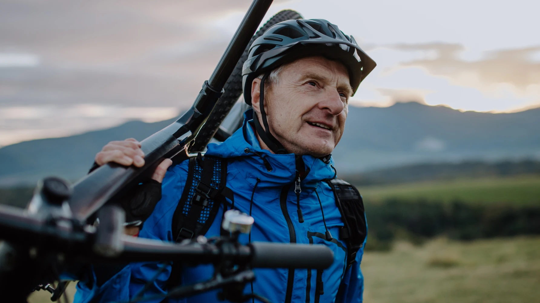 An older man wearing a blue jacket and black bicycle helmet smiles while carrying a mountain bike over his shoulder. He stands in a scenic outdoor setting with green fields and distant mountains under a cloudy sky during sunset or sunrise.