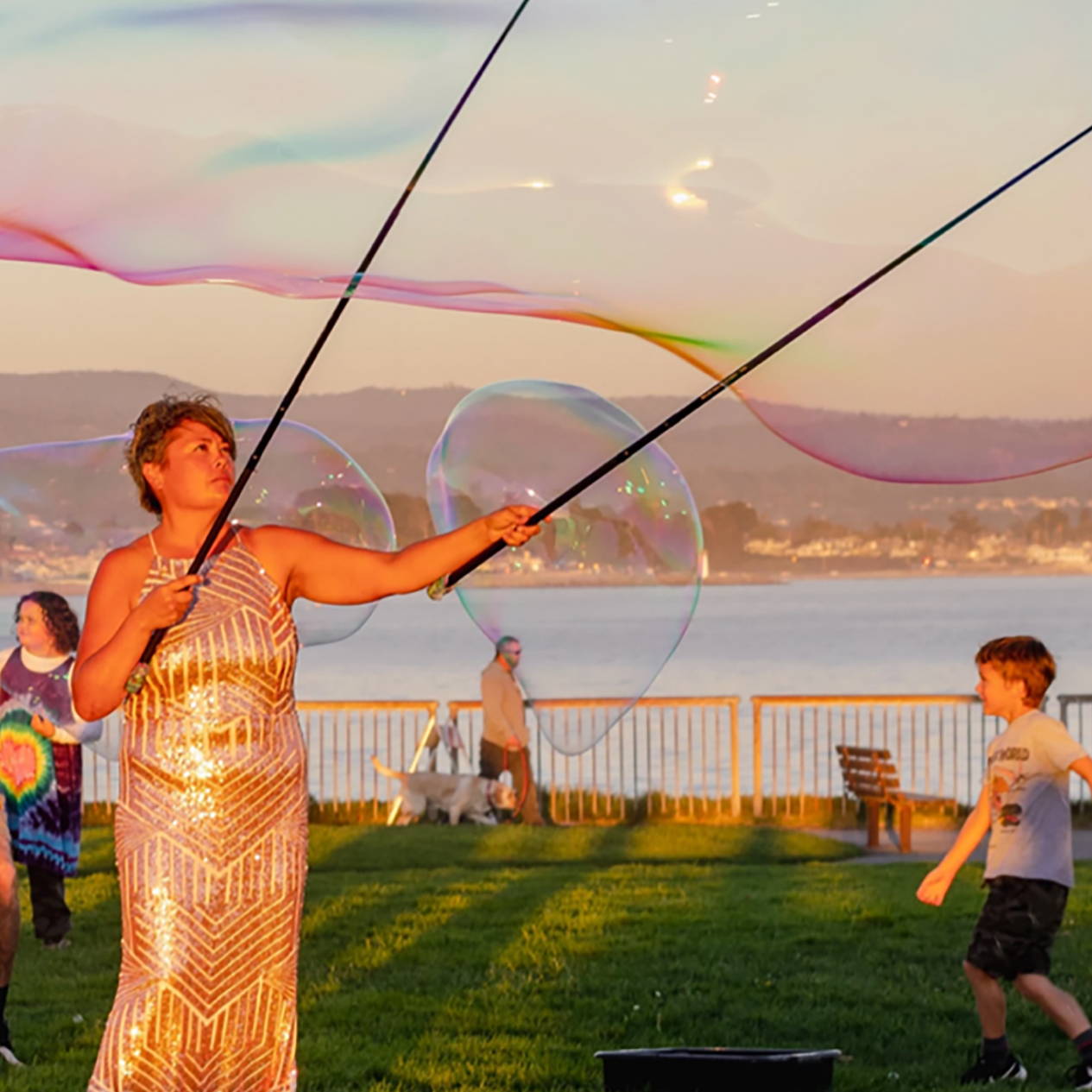 A woman in a sparkling dress creates giant iridescent soap bubbles with long wands at a park near a waterfront during sunset. Children play and adults watch and take photos, with scenic mountains and calm water in the background.